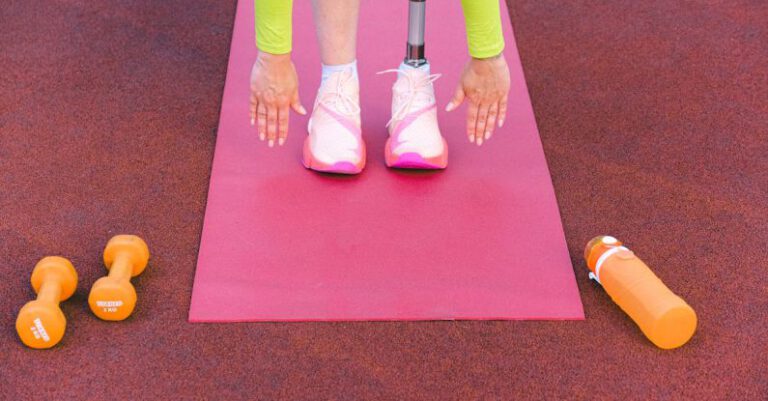 Stamina - Unrecognizable female athlete with leg prosthesis bending forward and stretching while trying ti touch ground during training on sports ground with dumbbells