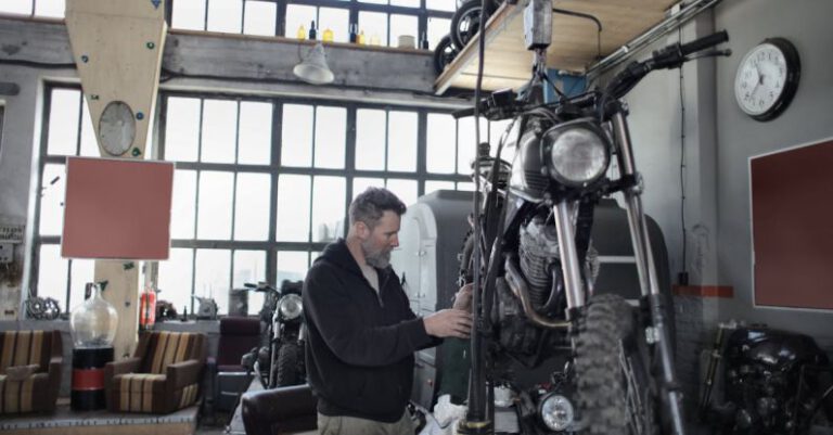 Bike Check - Side view of bearded adult mechanic in casual uniform standing near motorcycle and working with engine in workshop