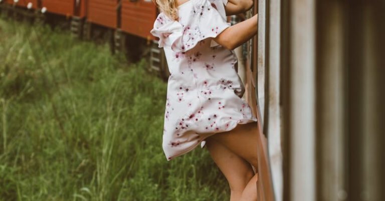 Road Hazards - Side view of full body young woman in air dress leaning out from exit door and holding on to railroad while riding train past green grass